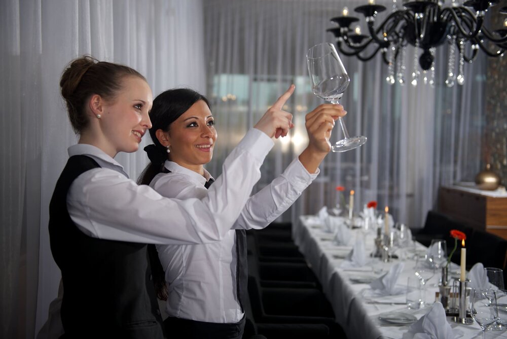 Zwei Mitarbeiterinnen stehen vor einer festlich gedeckten Tafel. Eine hält ein Weingals in die Höhe gegen das Licht, während die andere mit dem Finger darauf deutet.