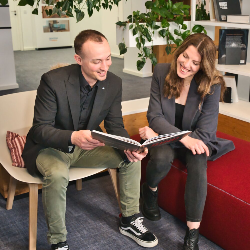 Ein Mann und eine Frau sitzen über Eck auf einem Stuhl und einer roten Bank. Sie lachen und schauen zusammen in ein Buch. 