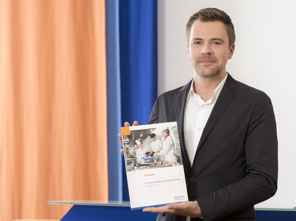 Lächelnder Mann in Anzug hält eine Mappe der DEHOGA Akadmie in die Kamer