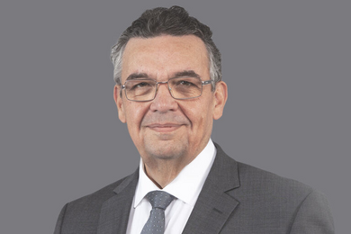 Porträt dunkelhaariger, lächelnder Mann mit Brille in Anzug und Krawatte. 