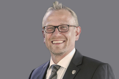 Porträt grauhaariger, lächelnder Mann mit Brille in Anzug und Krawatte.