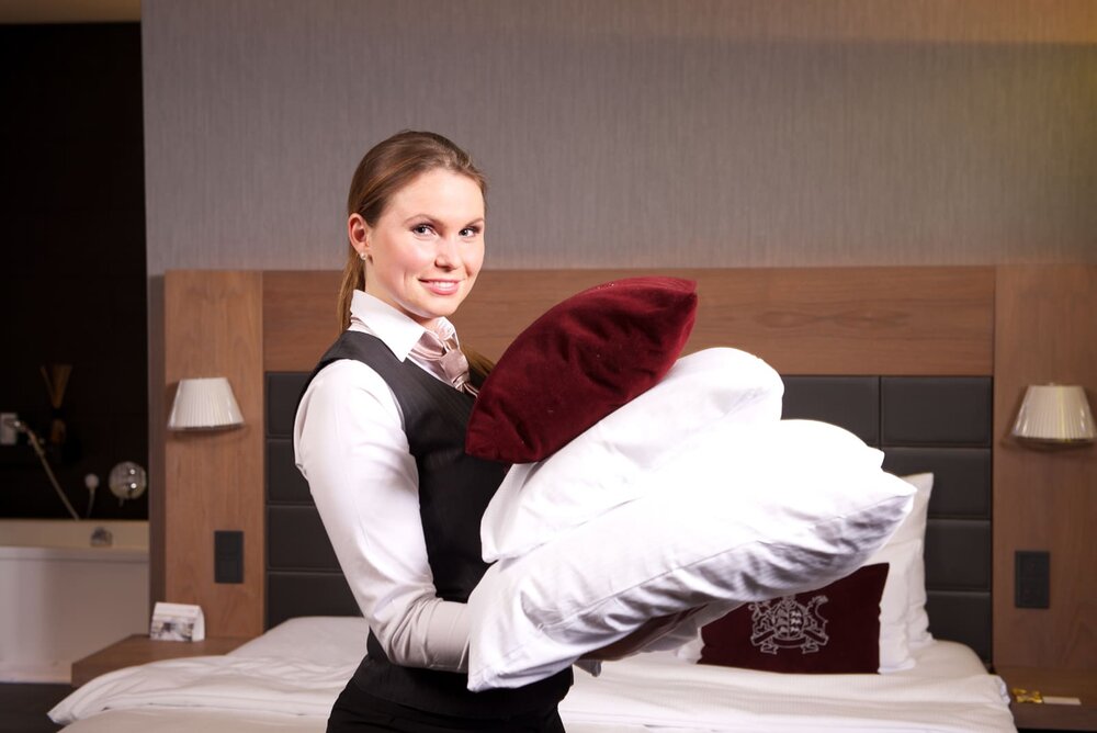 Eine Housekeeping-Mitarbeiterin in Hemd und Weste trägt Kissen und lächelt dabei in die Kamera