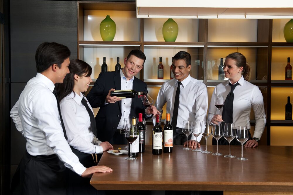 Ein Sommelier und vier Teilnehmer:innenstehen um einen Tisch auf dem verschiedene Weinflaschen und leere Weingläser stehen. Die Teilnehmer:innen tragen ein weißes Hemd mit Krawatte und lächeln. Der Sommelier schenkt Wein ein.