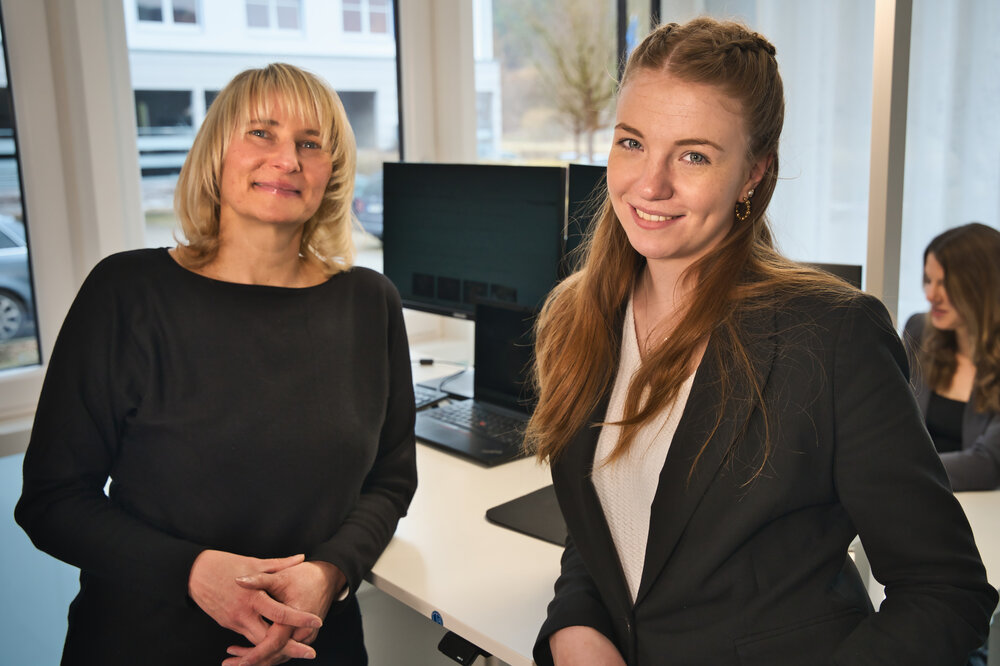 Zwei Frauen lächeln in die Kamera. Sie stehen in einem Büro. 