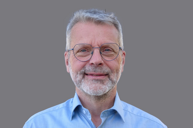Porträt: Grauhaariger, lächelnder Mann mit Brille. Blaues Hemd. 