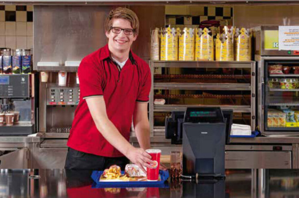 Lachender Mitarbeiter ein Fast-Food-Kette, der gerade ein Getränk auf ein Tablett mit Burger und Pommes stellt. 