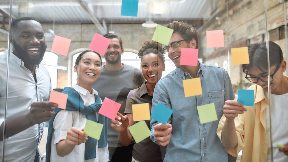 Blick durch eine Glasscheibe die mit bunten Post It's beklebt sind. Dahinter eine Gruppe von lachenden Menschen