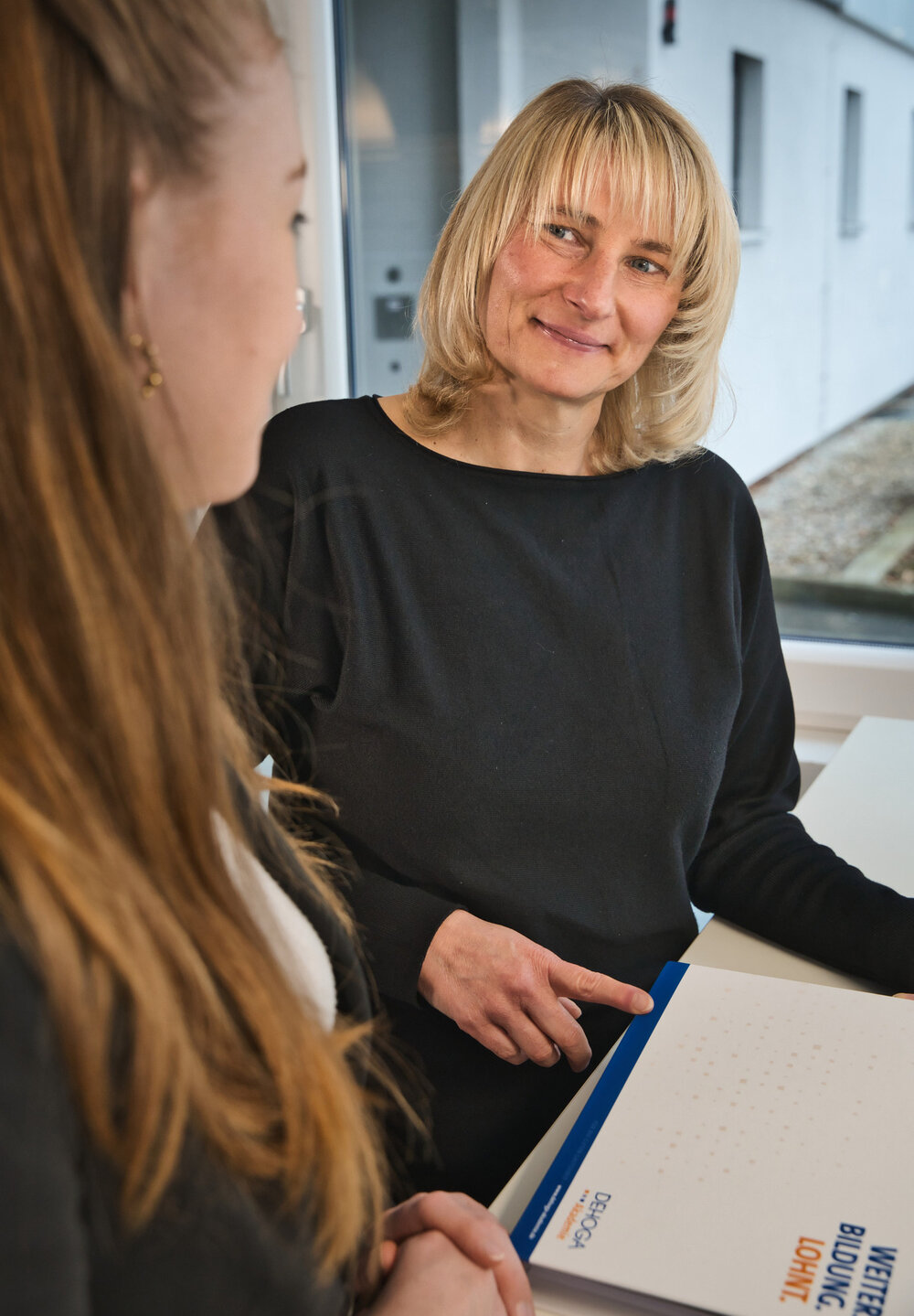 Zwei Frauen, eine verschwommen im Vordergrund. Die zweite Frau -blond - deutet lächelnd auf eine Mappe mit dem Titel ,,Weiterbildung lohnt''.