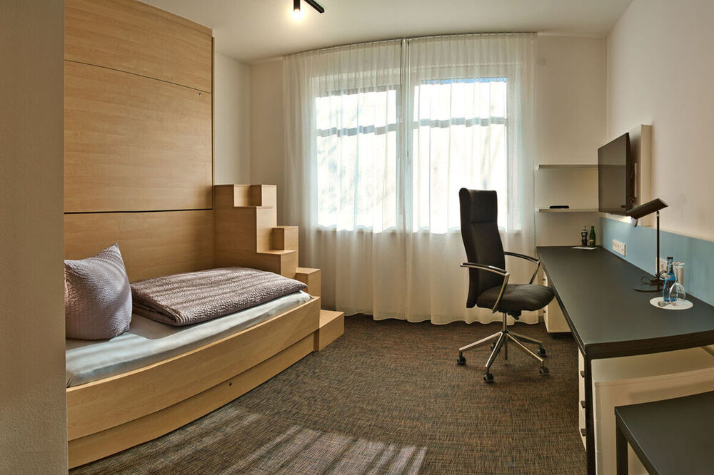 Zimmer. Links im Bild: gemachtes Einzelbett. Rechts: Schreibtisch mit Stuhl, Lampe und TV an der Wand.