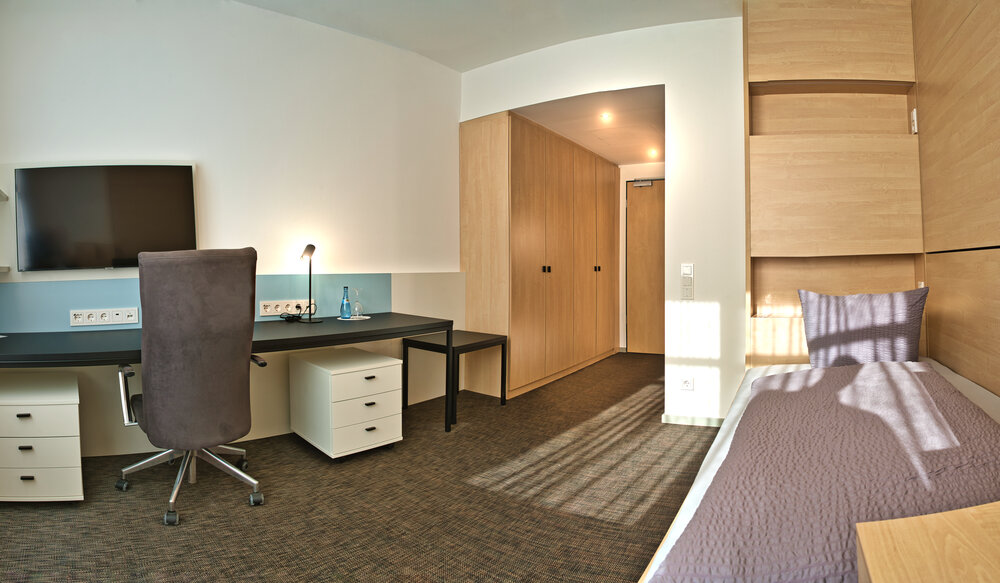 Blick in ein Zimmer. Links im Bild: Schreibtisch mit Stuhl, TV an der Wand. Rechts im Bild: Gemachtes Einzelbett. 