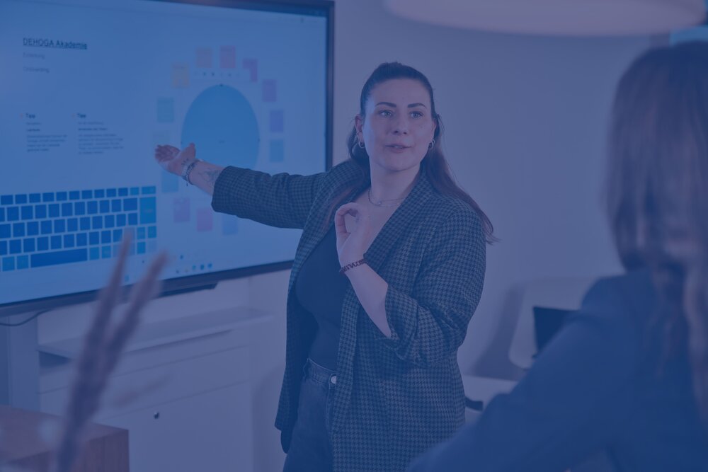 Blau eingefärbtes Bild: Eine Frau steht an einem Whiteboard und deutet darauf. Im vordergrund verschwommen eine weitere Person, zu der sie spricht.