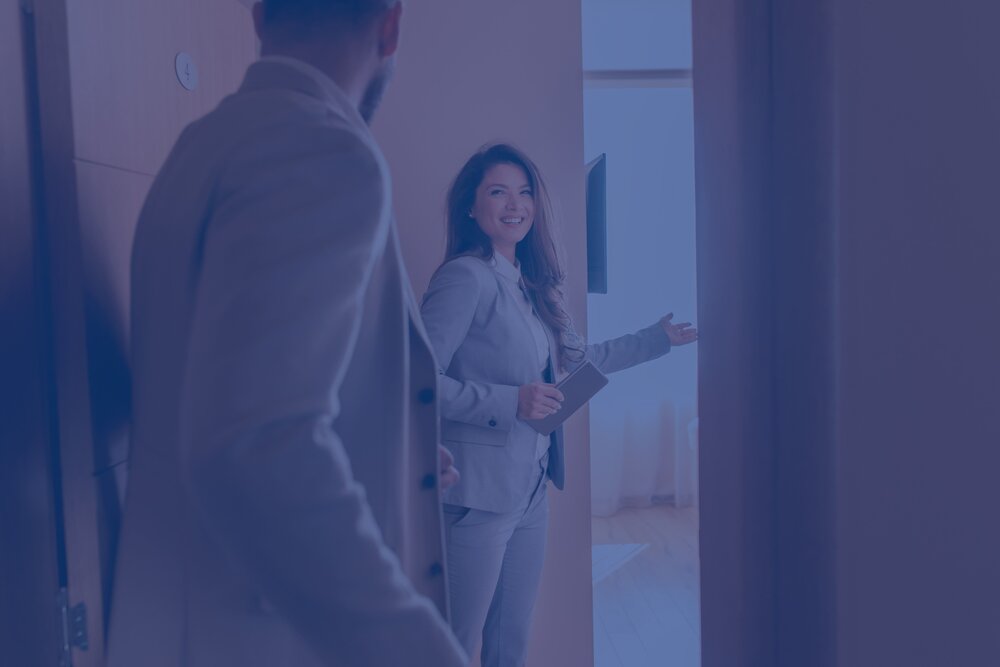 Blau eingefärbtes Bild: Zwei Menschen betreten ein Zimmer, eine Dame steht in der Tür und weist den Weg hinein