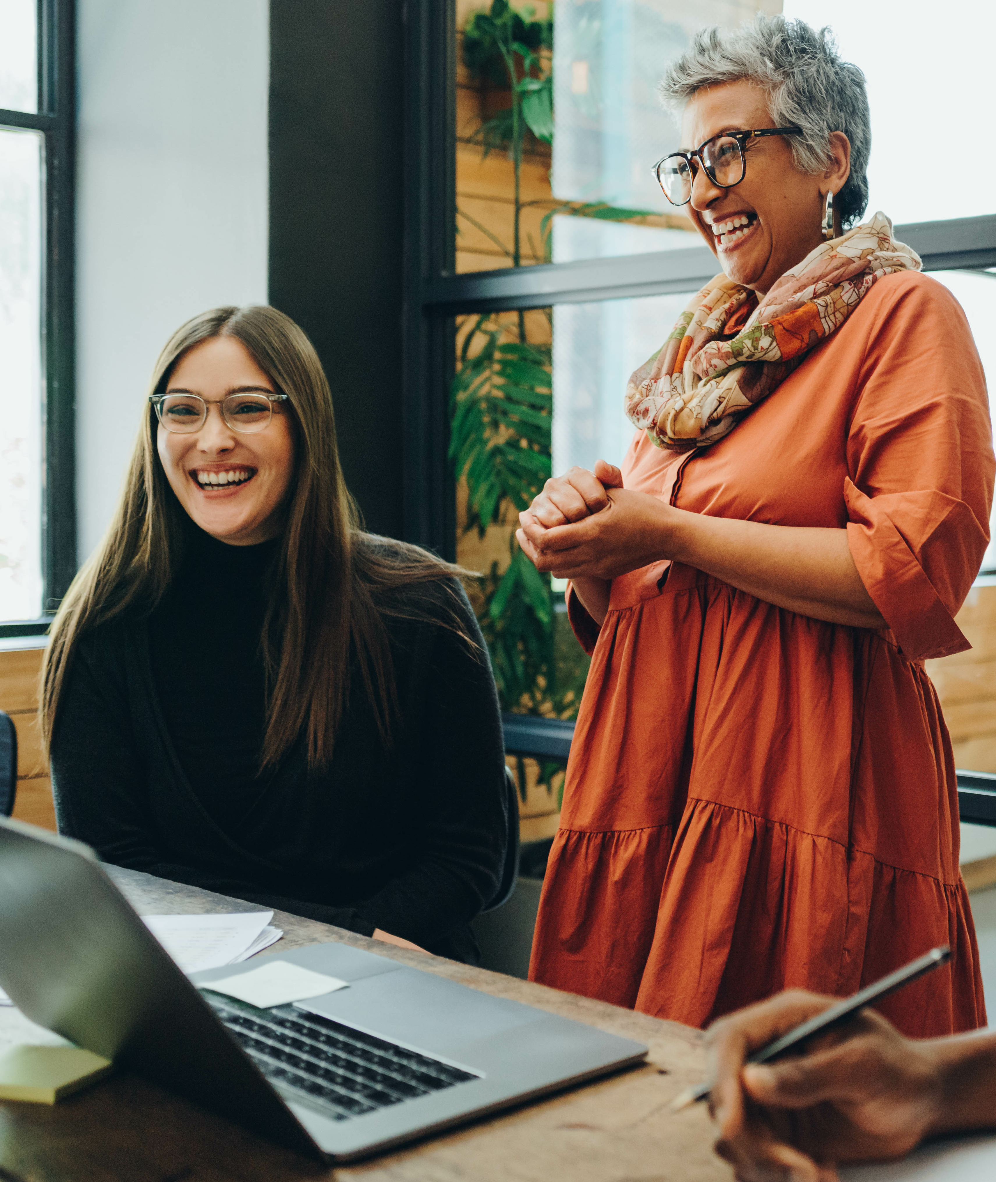Zwei lachende Frauen, eine steht eine sitzt an einem Tisch. Auf dem Tische in aufgeklappter Laptop