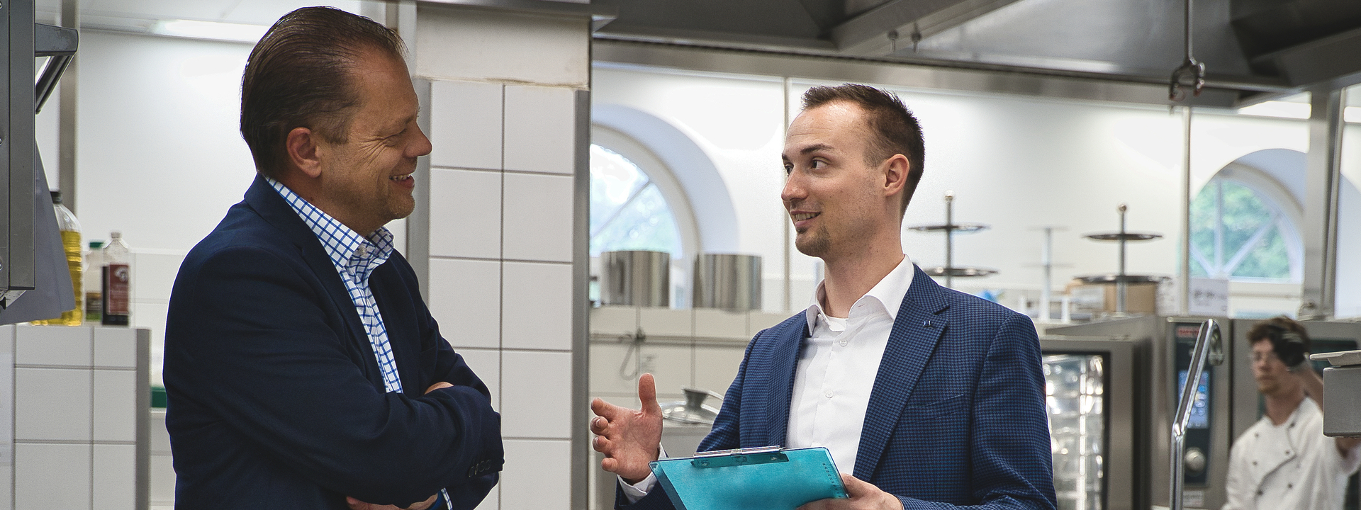 Zwei Männer im Anzug stehen in einer Küche und schauen sich an. Der rechte Mann erklärt dem linken lächelnd etwas. Er hält ein iPad in der Hand