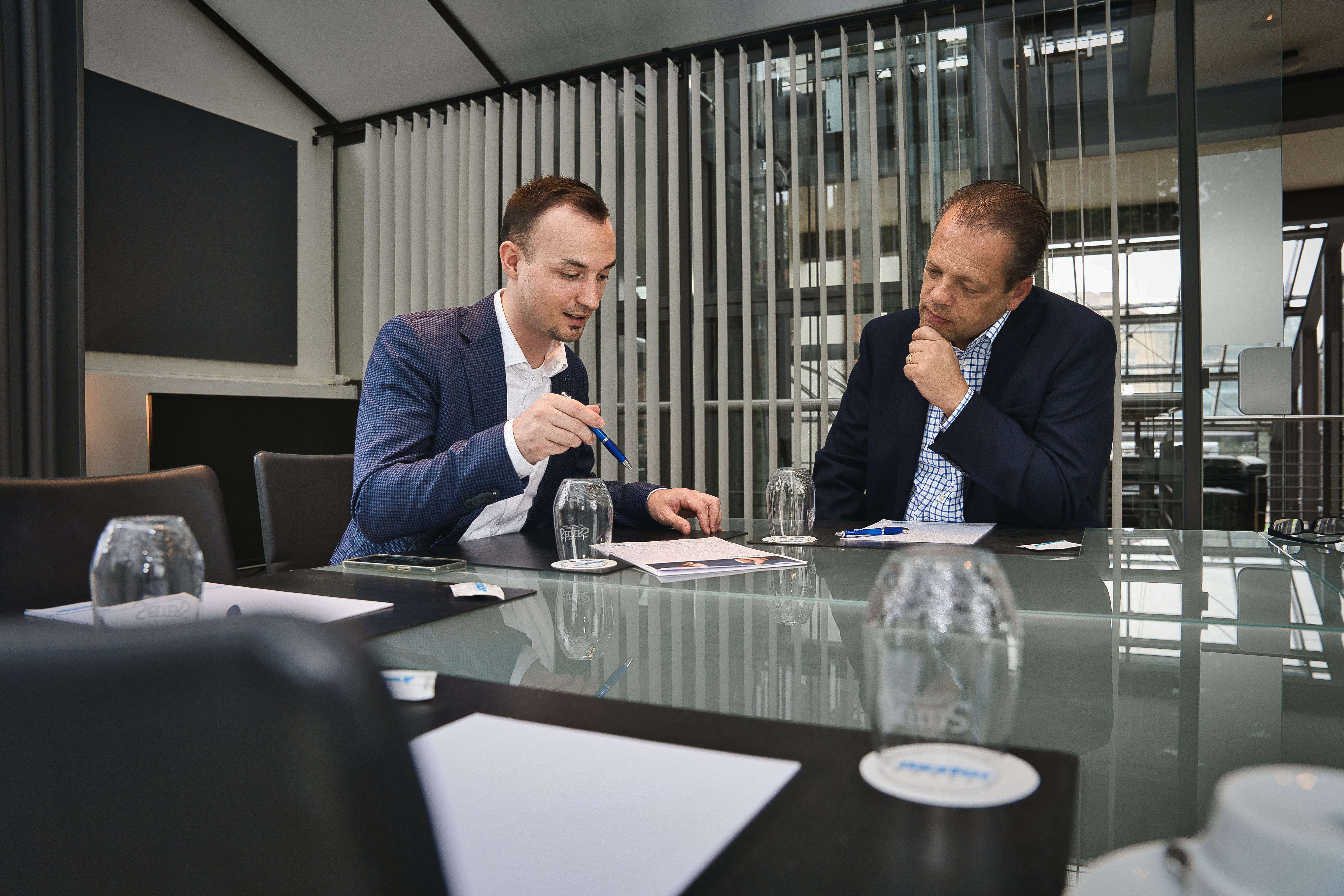 Zwei Männer sitzen an einem Schreibtisch. Der linke Mann deutet mit einem Stift auf ein Blatt Papier während der rechte zuhört